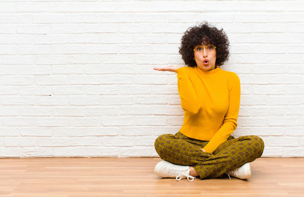 young pretty afro woman looking surprised and shocked, with jaw dropped holding an object with an open hand on the side sitting on the floor - Photo, Image