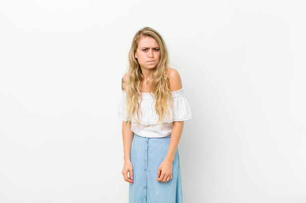 young blonde woman with a goofy, crazy, surprised expression, puffing cheeks, feeling stuffed, fat and full of food against white wall - Foto, afbeelding