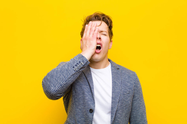 young red head man looking sleepy, bored and yawning, with a headache and one hand covering half the face against orange background - 写真・画像