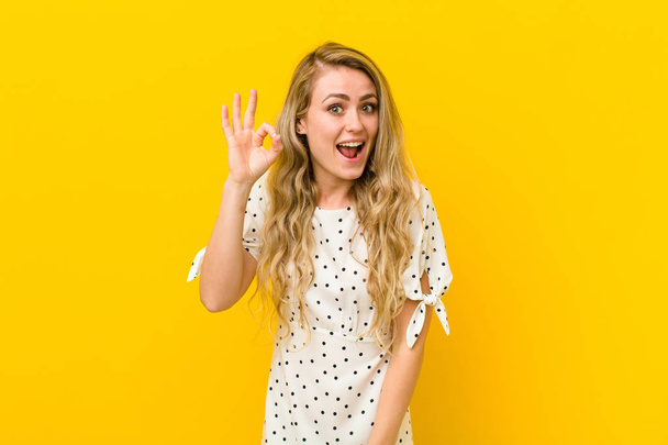 young blonde woman feeling successful and satisfied, smiling with mouth wide open, making okay sign with hand against yellow wall - Photo, Image