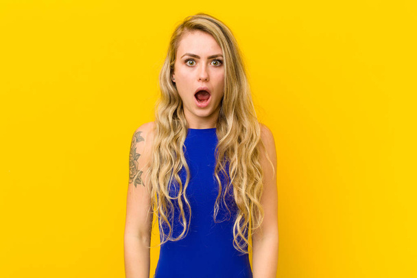 young blonde woman looking shocked, angry, annoyed or disappointed, open mouthed and furious against yellow wall - Photo, Image