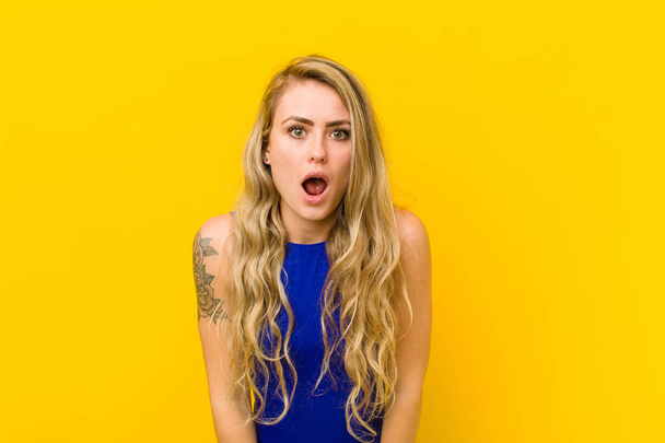 young blonde woman shouting aggressively, looking very angry, frustrated, outraged or annoyed, screaming no against yellow wall - Zdjęcie, obraz