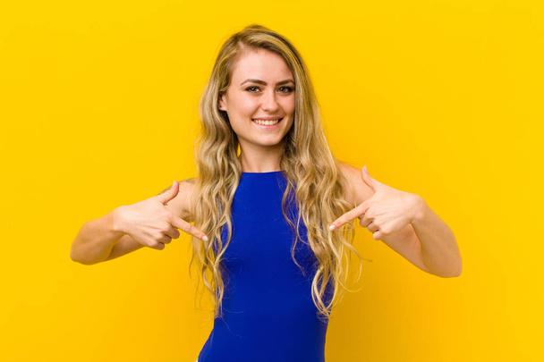 young blonde woman looking proud, arrogant, happy, surprised and satisfied, pointing to self, feeling like a winner against yellow wall - Foto, immagini