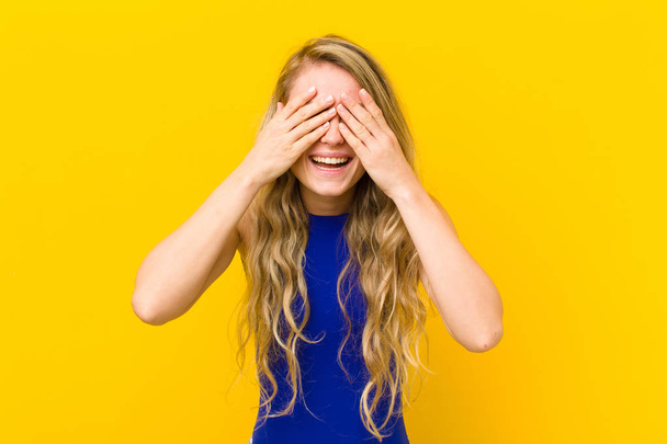 young blonde woman smiling and feeling happy, covering eyes with both hands and waiting for unbelievable surprise against yellow wall - Fotografie, Obrázek