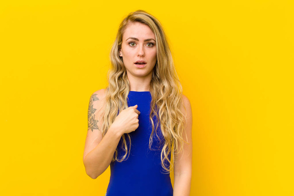 young blonde woman looking happy, proud and surprised, cheerfully pointing to self, feeling confident and lofty against yellow wall - Foto, afbeelding