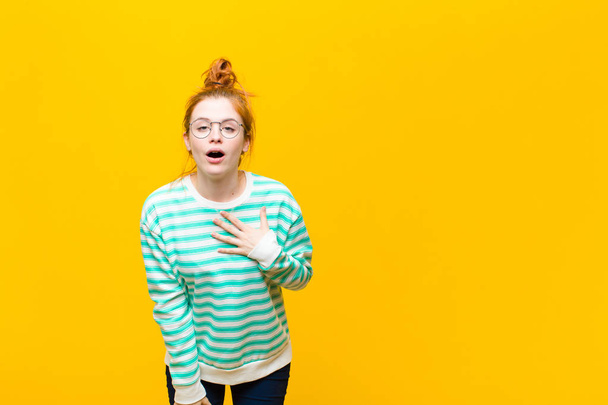 young red head woman feeling shocked and surprised, smiling, taking hand to heart, happy to be the one or showing gratitude against orange wall - Foto, Imagen
