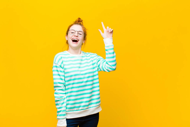 young red head woman feeling like a happy and excited genius after realizing an idea, cheerfully raising finger, eureka! against orange wall - Foto, imagen
