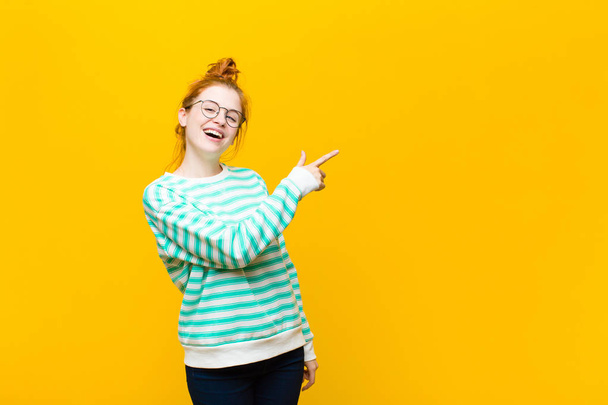 young red head woman looking excited and surprised pointing to the side and upwards to copy space against orange wall - Foto, afbeelding