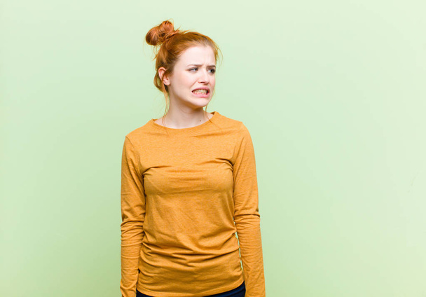young pretty red head woman looking worried, stressed, anxious and scared, panicking and clenching teeth against green wall - Foto, Imagen