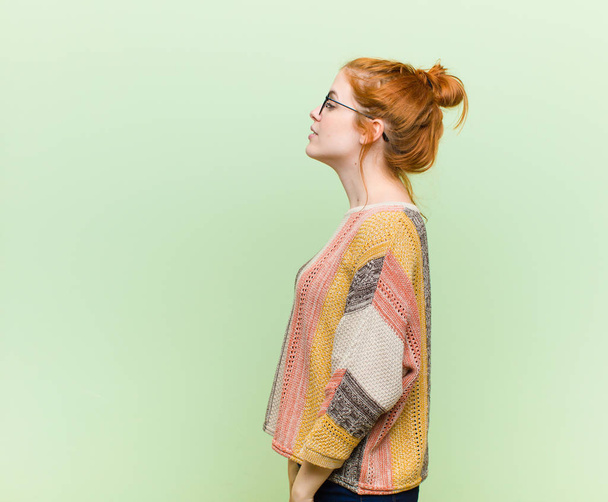 young pretty red head woman on profile view looking to copy space ahead, thinking, imagining or daydreaming against green wall - Fotoğraf, Görsel