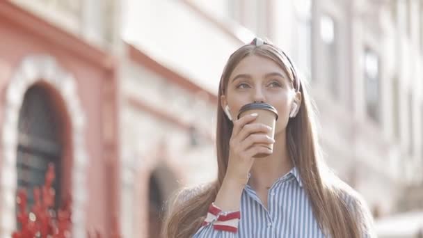 Portrait of Cheerful Smiling Young Pretty Girl Wearing Stylish Headband and Striped Shirt in Earphones, Drinking Coffee, Walking Happily and Looking Around at The Old City Background. - Πλάνα, βίντεο