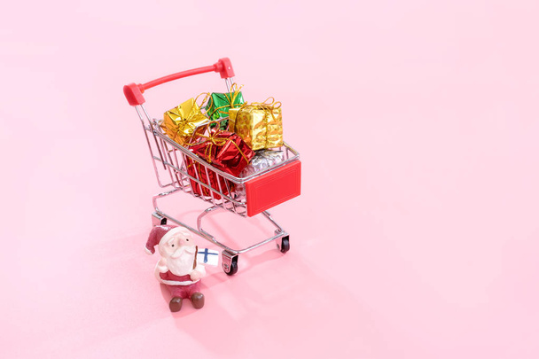 Christmas shopping concept, mini red shop cart trolley with Santa Claus toy and gift box isolated on pale pink background, blank copy space, close up - Fotoğraf, Görsel