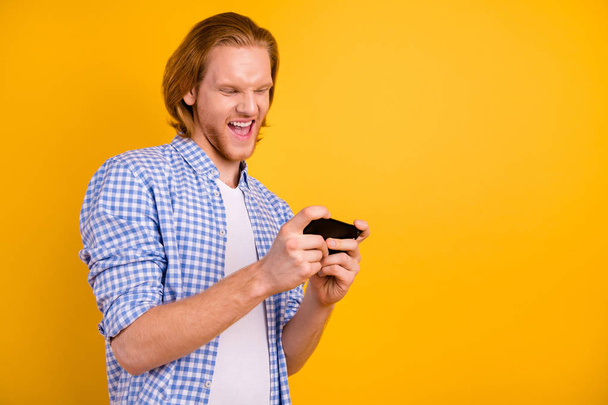 Photo of trendy casual cheerful playing hipster creating copyspace holding mobile phone with hands wearing blue shirt isolated over yellow vivid color background - Φωτογραφία, εικόνα