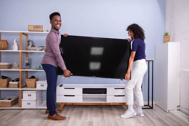 Side View Of Young Couple Lifting Television At Home - Foto, afbeelding