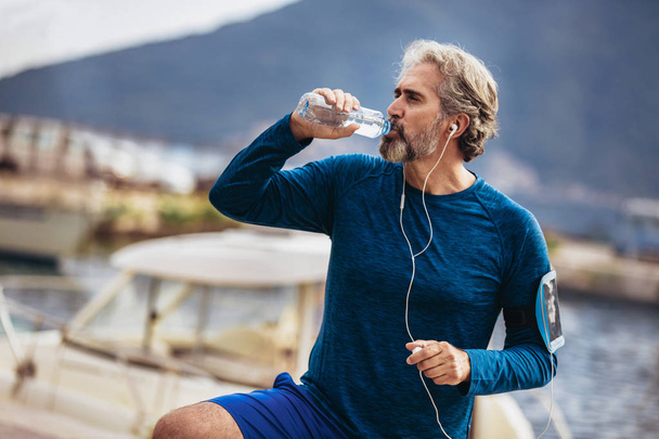 Fit thirsty senior man drinking water before running. Active old - Photo, Image