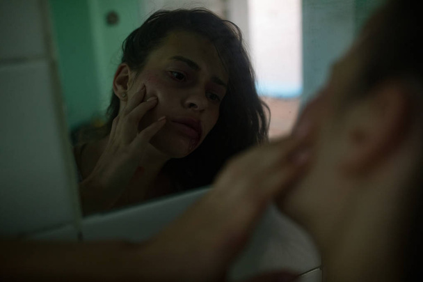Избитая окровавленная женщина-жертва насилия исследует свои раны на лице в зеркале
.  - Фото, изображение