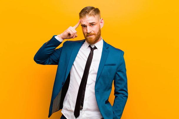 young red head businessman feeling confused and puzzled, showing you are insane, crazy or out of your mind against orange background - 写真・画像