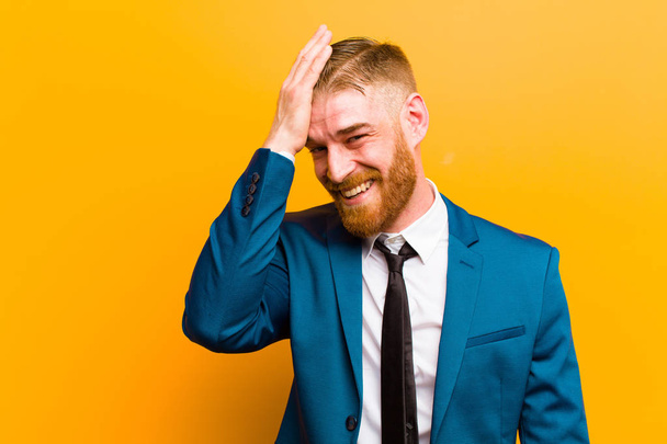 young red head businessman laughing and slapping forehead like saying doh! I forgot or that was a stupid mistake against orange background  - Fotó, kép