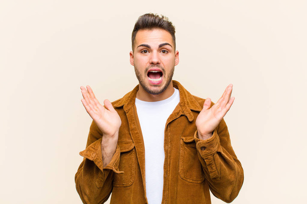young hispanic man looking happy and excited, shocked with an unexpected surprise with both hands open next to face against isolated background - Photo, Image