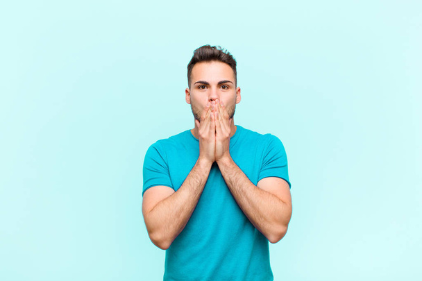 young hispanic man happy and excited, surprised and amazed covering mouth with hands, giggling with a cute expression against blue background - Foto, imagen
