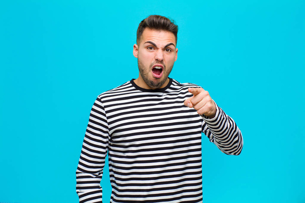 young hispanic man pointing at camera with an angry aggressive expression looking like a furious, crazy boss against blue background - Zdjęcie, obraz