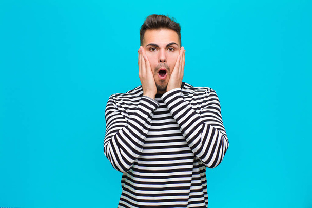 young hispanic man feeling shocked and scared, looking terrified with open mouth and hands on cheeks against blue background - Photo, image