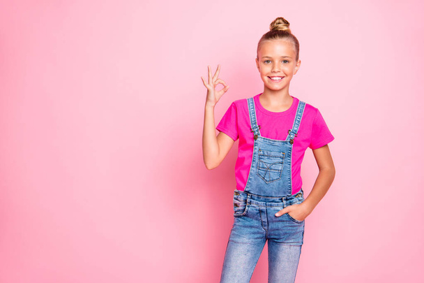 Photo of cheerful cute nice preteen girl showing you ok sign approving your actions holding hand in pocket of jeans overall isolated over pink pastel color background - Photo, Image