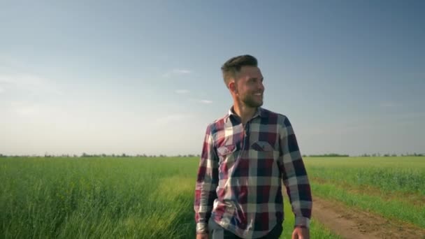 smiling male farmer enjoy the fresh air and the beauty of nature, young man goes to camera on background of green field and blue sky - Záběry, video