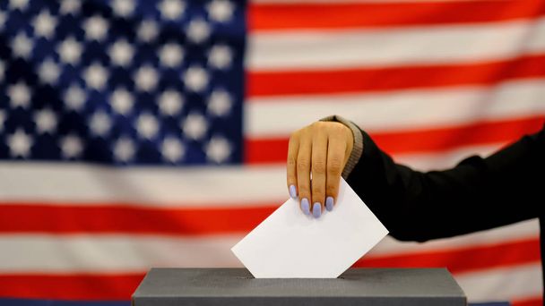 Kobieta oddanie głosowanie w polu głosowania w dniu wyborów. Bliska ręka z białym papierze głosowym na tle flagi USA. - Zdjęcie, obraz
