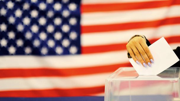 Eine Frau wirft am Wahltag einen Stimmzettel in eine Wahlurne. Nahaufnahme der Hand mit weißem Stimmzettel auf dem Hintergrund einer US-Flagge. - Foto, Bild