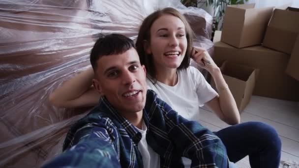 Excited young caucasian couple sitting on floor among cardboard boxes in new home and having video call with friends on smartphone, talking excitedly, smiling and gesturing - Кадры, видео