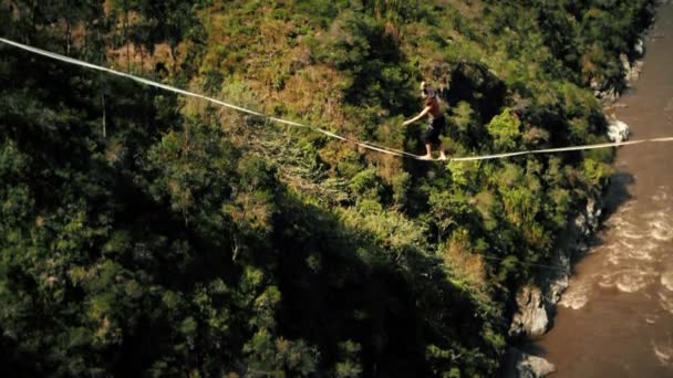 Jonge man lopen op slackline Tightrope tussen rotsen hoog boven de grond met groene bos en stromende rivier op de achtergrond - Video