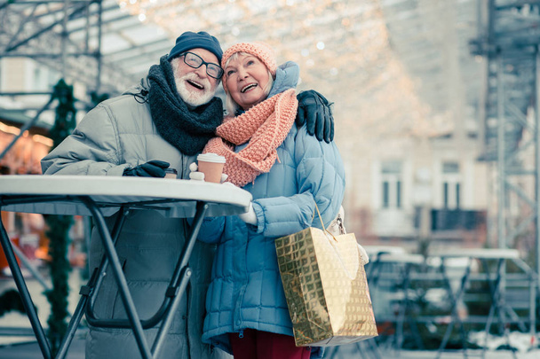 Positive aged woman with a shiny shopping bag leaning to her husband and smiling while standing outdoors near the round table with him - Photo, Image