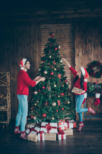 Vertical full length body size view of nice attractive cheerful cheery married spouses spending chistmastime having fun putting balls toys on fir in decorated loft industrial brick style interior - Foto, Imagem