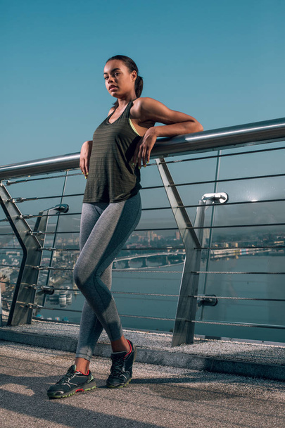 Confident fit sportswoman standing on the bridge with her elbows on the banister - 写真・画像