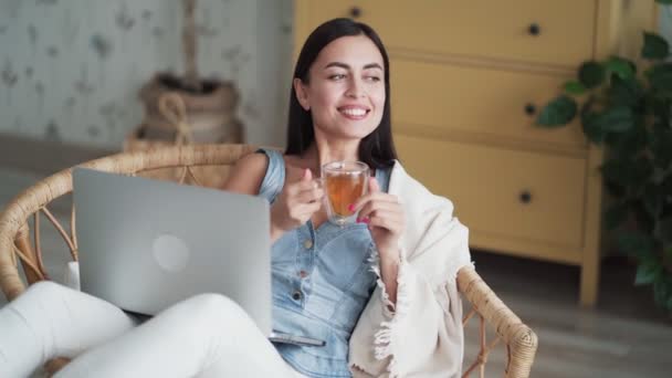 Woman sits in chair with laptop, holds cup of tea in her hands, drinks it, smiles - Séquence, vidéo