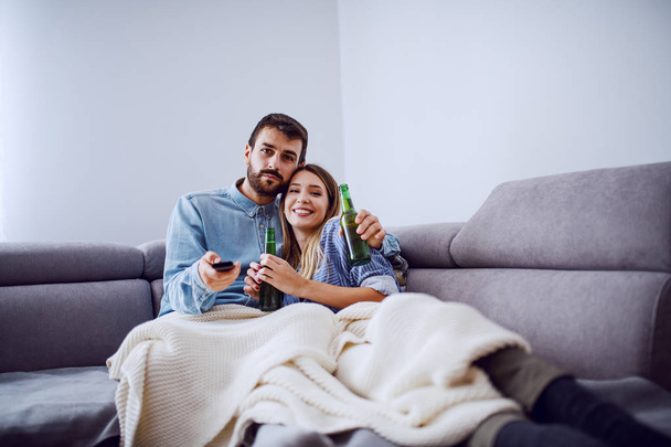 glückliches nettes kaukasisches Paar, das auf dem Sofa im Wohnzimmer sitzt, Bier trinkt und Film guckt. - Foto, Bild