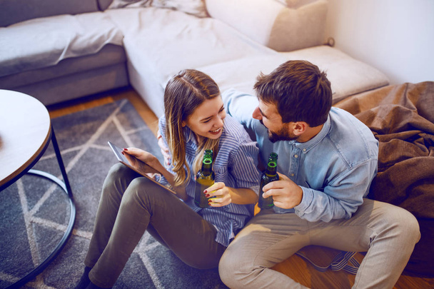 Attractive caucasian woman sitting on the floor and holding tablet and beer while looking at her boyfriend. Man holding beer, looking and hugging woman. - Photo, image