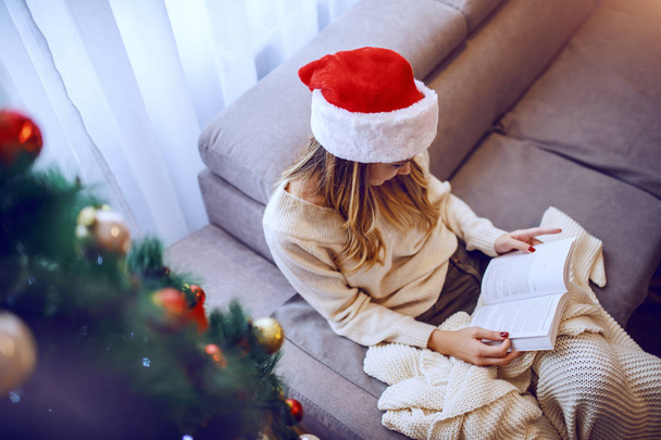 Widok z góry pięknej białej młodej kobiety w swetrze i kapelusz Mikołaja na głowie siedzi na kanapie obok choinki i książki. - Zdjęcie, obraz