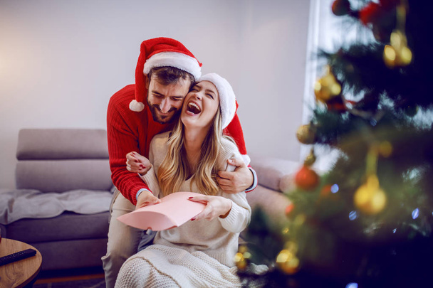 Handsome caucasian man giving christmas present to his loving girlfriend. Both are dressed in sweaters and having santa hat on heads. Christmas holidays. - Fotoğraf, Görsel