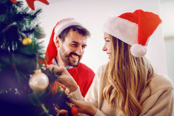 glückliches kaukasisches junges Paar mit Weihnachtsmützen auf dem Kopf, die den Weihnachtsbaum schmücken, während sie im Wohnzimmer stehen. - Foto, Bild
