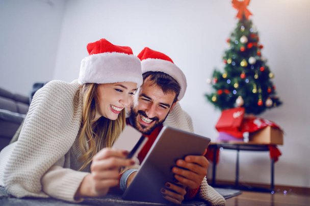Cute Caucasian couple lying on stomach on the floor with santa hats on heads and using tablet for online shopping. Woman holding credit card. Living room interior. In background is christmas tree. - 写真・画像