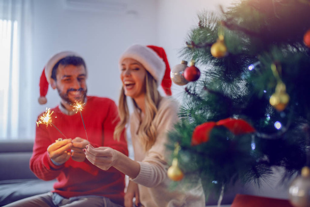 glückliches kaukasisches Paar mit Weihnachtsmannmützen auf dem Kopf, die Sprinkler in der Hand halten und Neujahr feiern. selektiver Fokus auf Sprinkler. Im Vordergrund steht der Weihnachtsbaum. - Foto, Bild