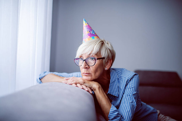 Verdrietig mooie blanke senior vrouw met bril en verjaardagskap op hoofd leunend op de bank en kijkend door het raam. Niemand herinnert zich haar verjaardag.. - Foto, afbeelding