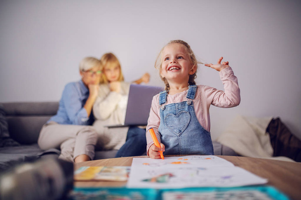 遊び心のある白人ブロンドの女の子は笑顔と絵を描きます.バックグラウンドで彼女のお母さんとおばあちゃんはノートパソコンを見て。リビングルームのインテリア。家族の時間. - 写真・画像