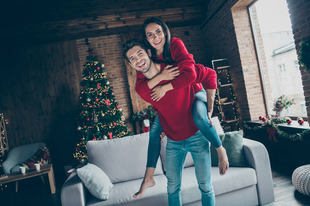 Portrait of romantic two youth hug piggyback on christmas x-mas party event wearing red jumper denim jeans in house with decoration illumination lights indoors near couch - Photo, Image
