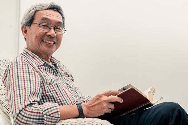 Evde sandalyede kitap okuyan Kıdemli Asyalı adam. - Fotoğraf, Görsel