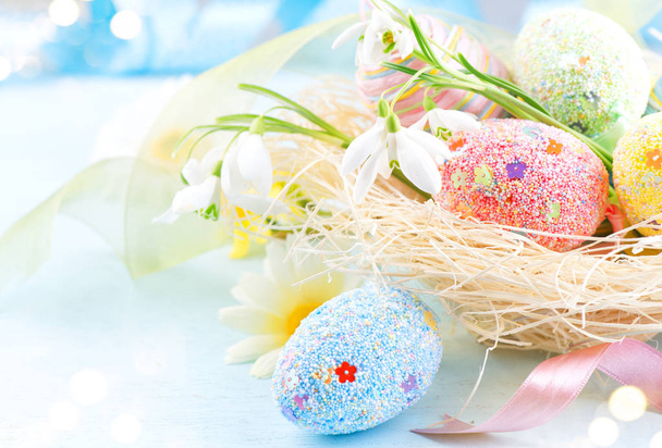 Πάσχα πολύχρωμα αυγά φόντο. Όμορφα πολύχρωμα αυγά με διακοσμητικά πάνω από μπλε ξύλινο φόντο, περίγραμμα του σχεδιασμού σε παστέλ χρώματα. Ανοιξιάτικα λουλούδια, κορδέλα διακοπών και τα αυγά του ζωγράφου στη φωλιά - Φωτογραφία, εικόνα