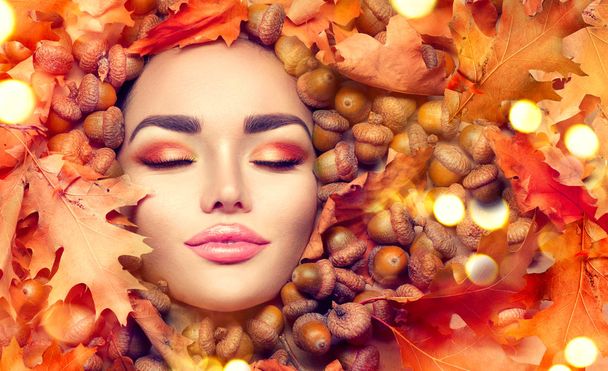 Herfst vrouw make-up. Mooie herfst model meisje gezicht portret met helder geel, rood en oranje kleur bladeren en eiken eikels - Foto, afbeelding