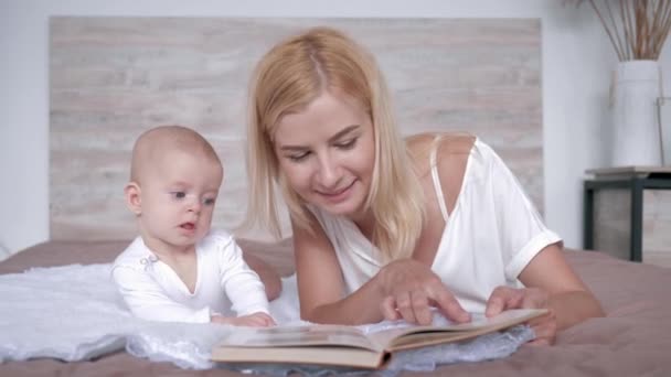 caring mom with book reads fairy tale story to her baby daughter then kisses her lying on bed at home - Filmmaterial, Video